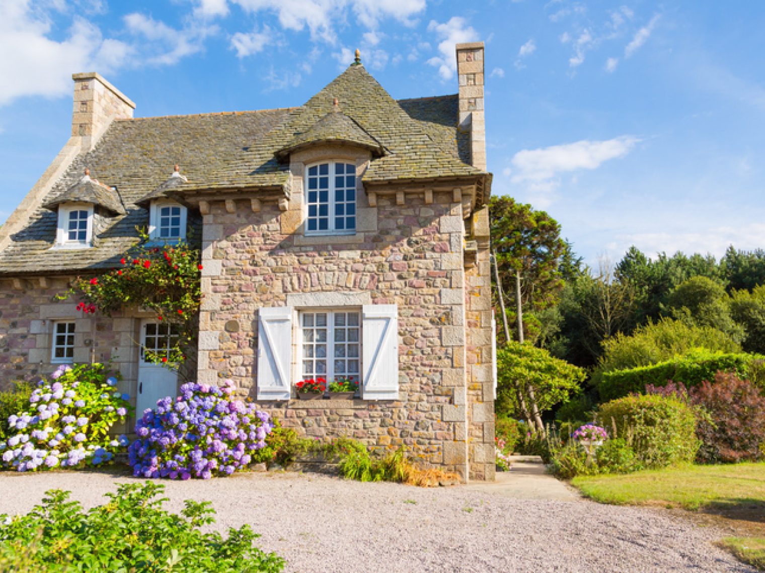 A charming stone cottage with a steep roof, adorned with colorful flowers and shutters, set in a lush garden under a blue sky.