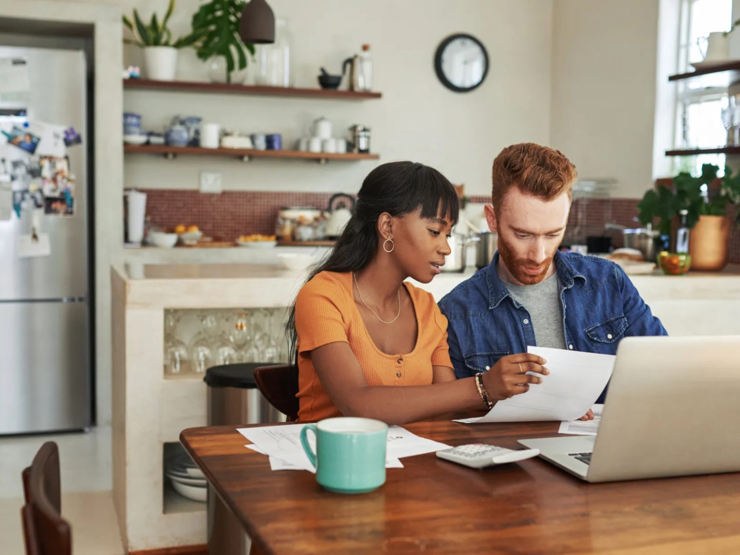 A man and woman sit at a kitchen table looking intently at a document together, with a laptop open in front of them.