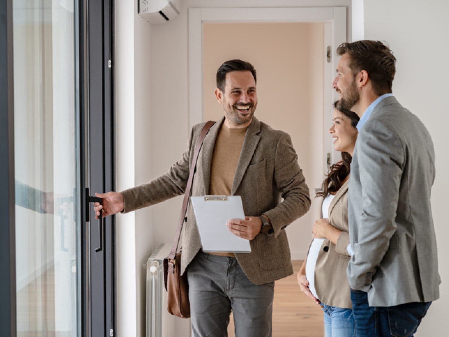 A real estate agent, holding documents, is smiling and gesturing welcomingly to a couple as they enter a home.