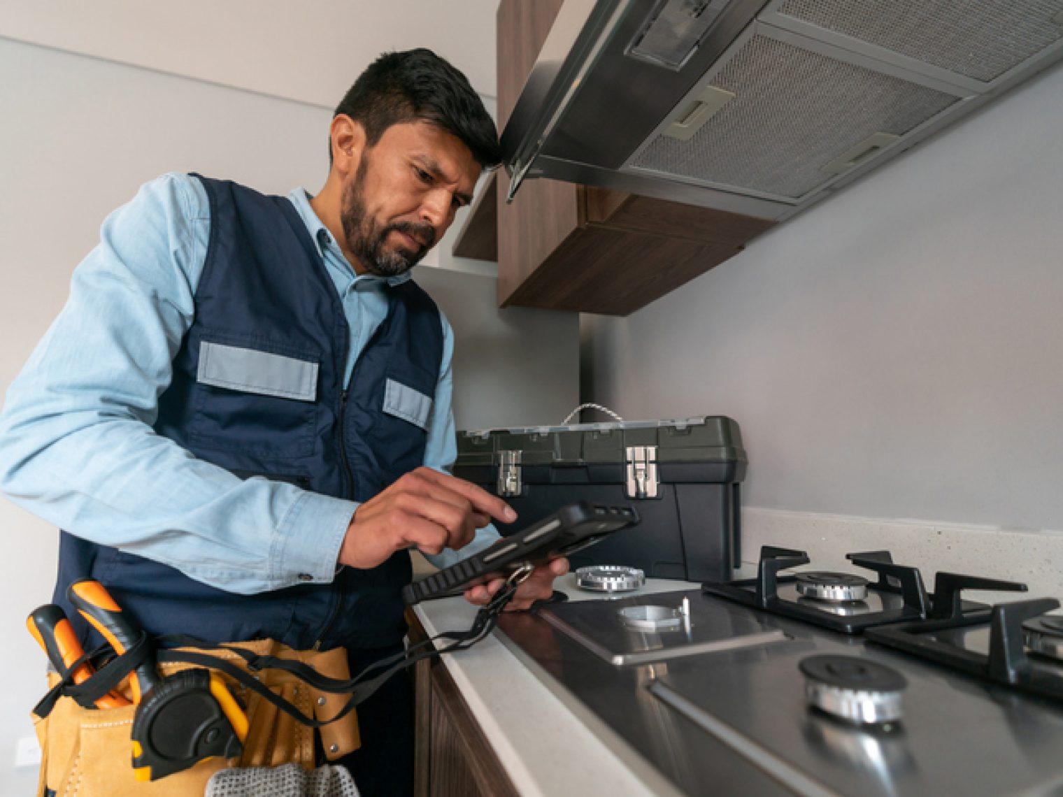 An inspector wearing a tool belt is examining a kitchen appliance with a handheld device.