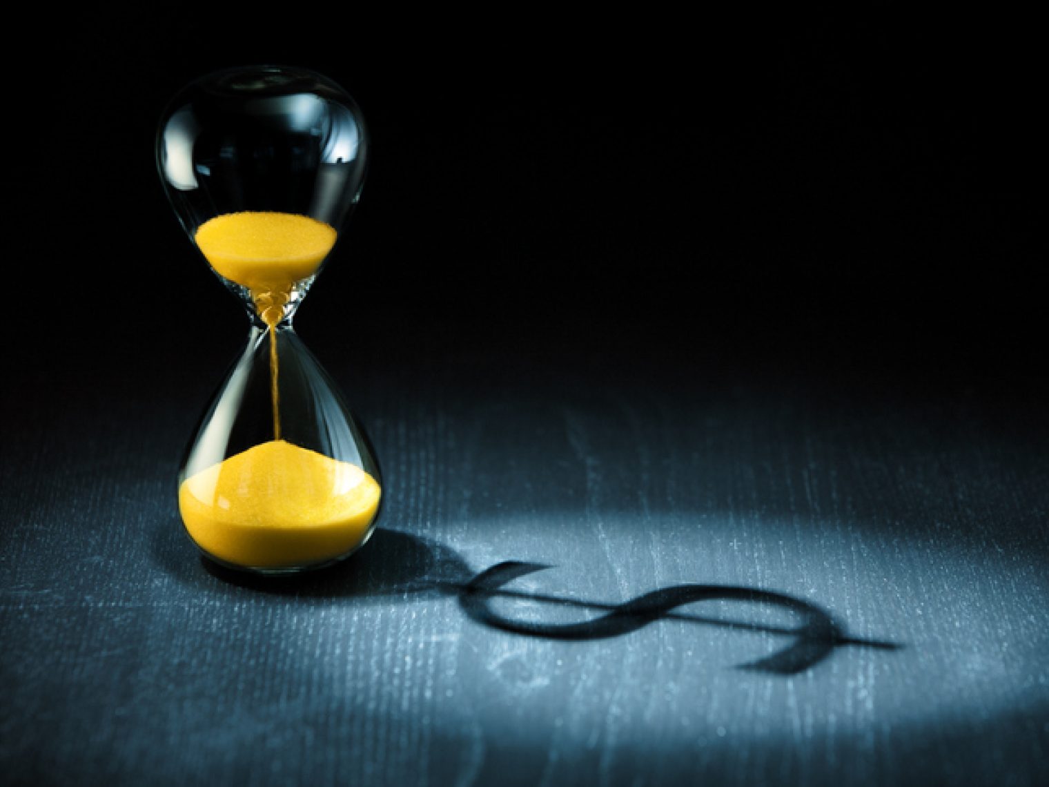 An hourglass with yellow sand on a dark background, casting a long shadow in the shape of a dollar sign.