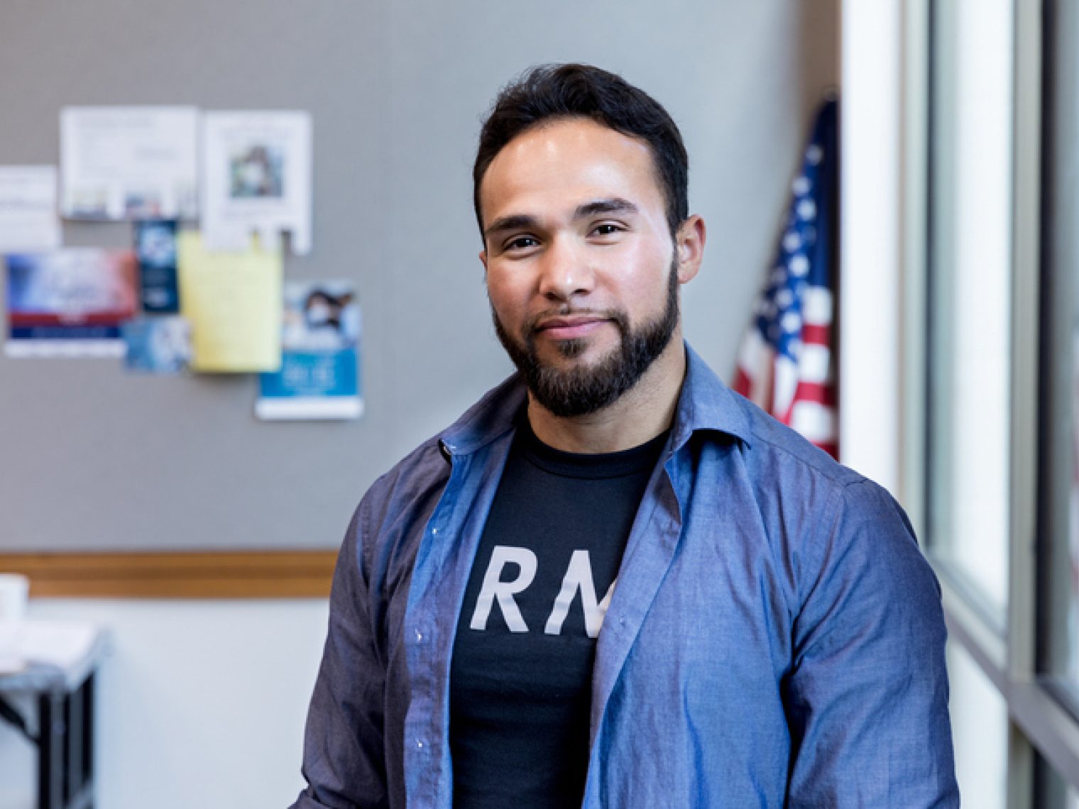 A confident man in a blue shirt over a graphic tee poses for a portrait, with an American flag in the background.