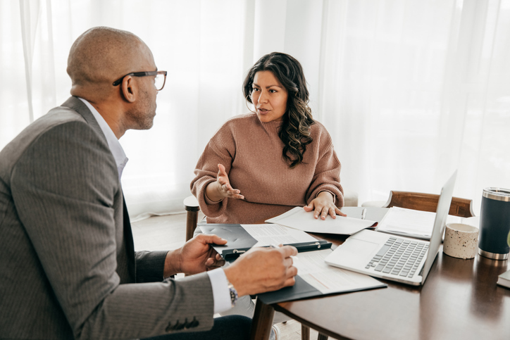 A focused conversation is taking place between a woman and a man sitting at a table with documents, a laptop, and a tablet, in what appears to be a serious professional consultation.