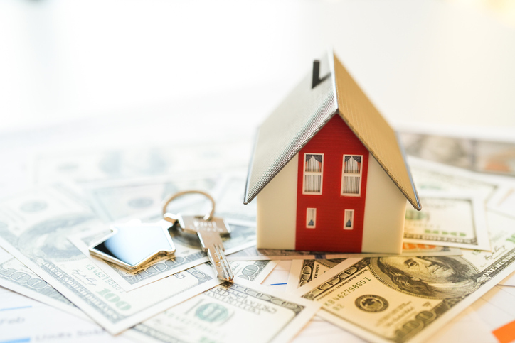 Miniature house on dollars on table with a pair of house keys to the side.