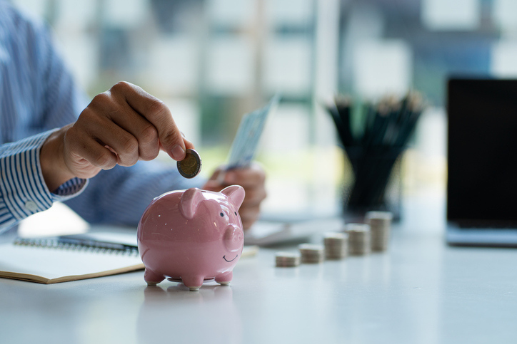 A person in a striped shirt is inserting a coin into a pink piggy bank, with a laptop and stacks of coins on a desk, symbolizing savings or financial planning.