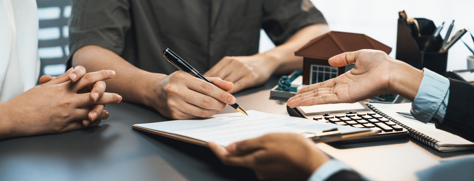 A close-up of a couple's hands on a desk as they are about to sign a document, with a real estate agent gesturing.