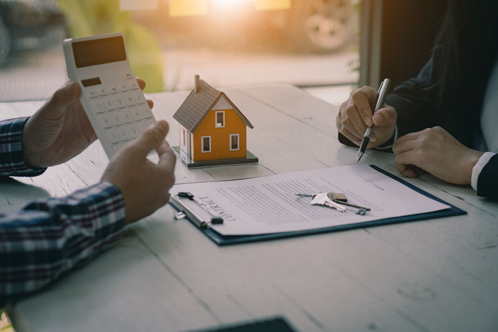 Hands of two people at a table with one holding a calculator and the other signing a document, with a small house model and keys nearby, suggesting a financial transaction or agreement.